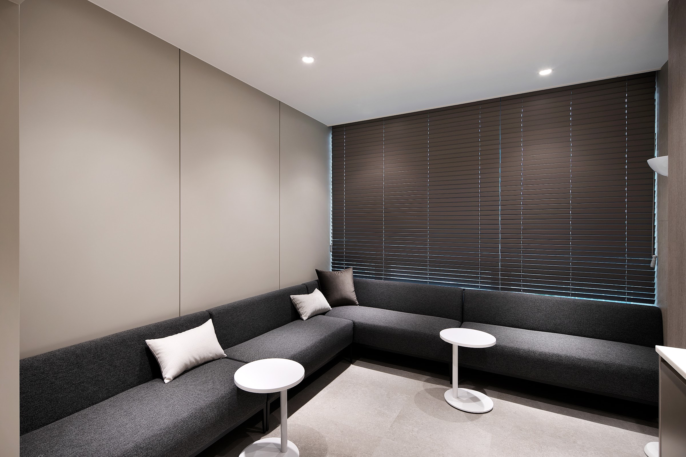 Quiet clinic lounge with a dark grey corner sofa, white cushions, small round white tables and lowered wooden blinds